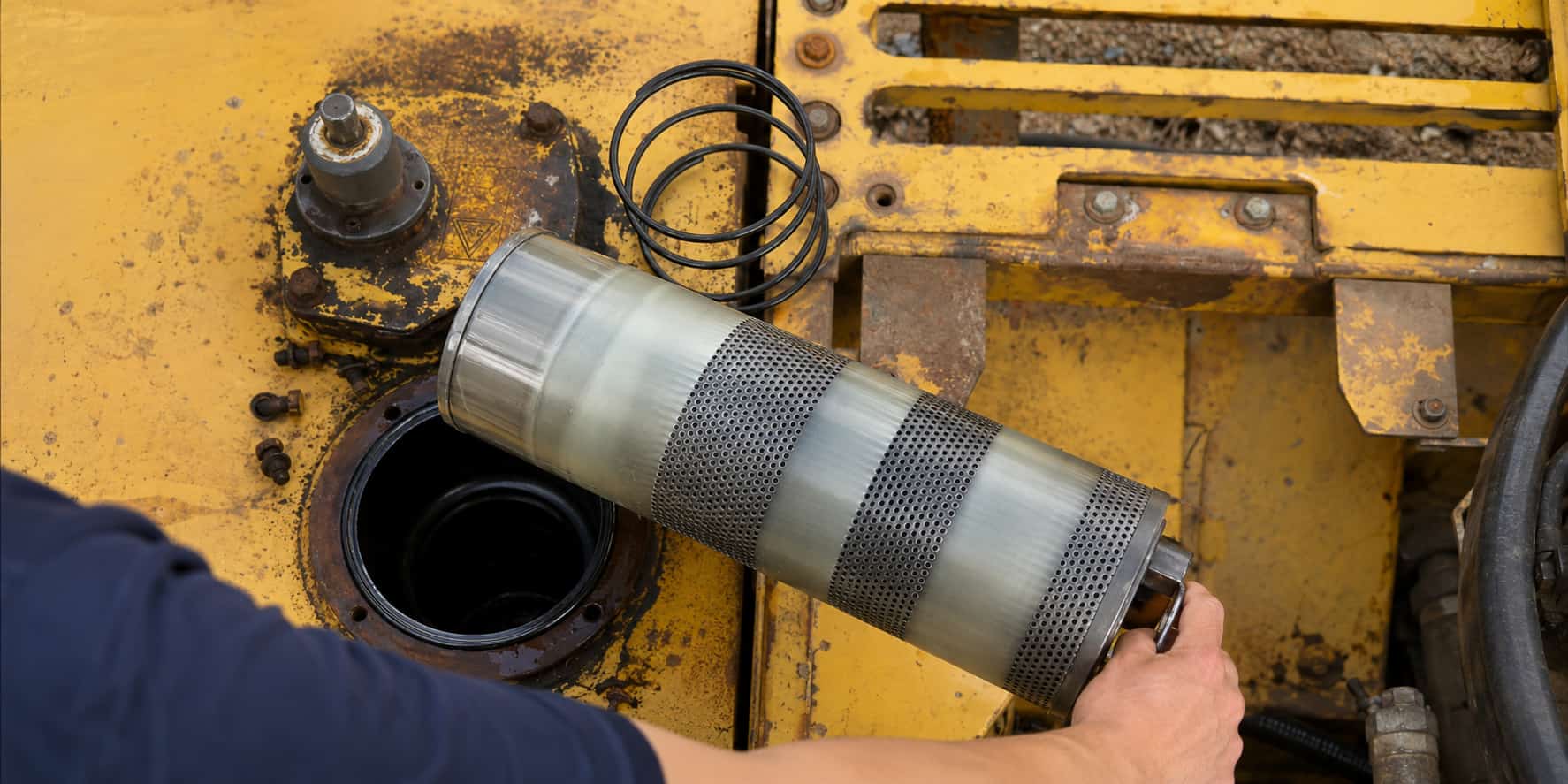 Excavator hydraulic return filter being removed from the machine during maintenance, showing the filter element and hydraulic tank opening